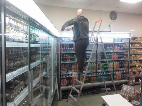 Mężczyzna na drabinie serwisuje chłodniczą witrynę sklepową z napojami, widoczne wentylatory i oświetlenie. Ujęcie od tyłu.