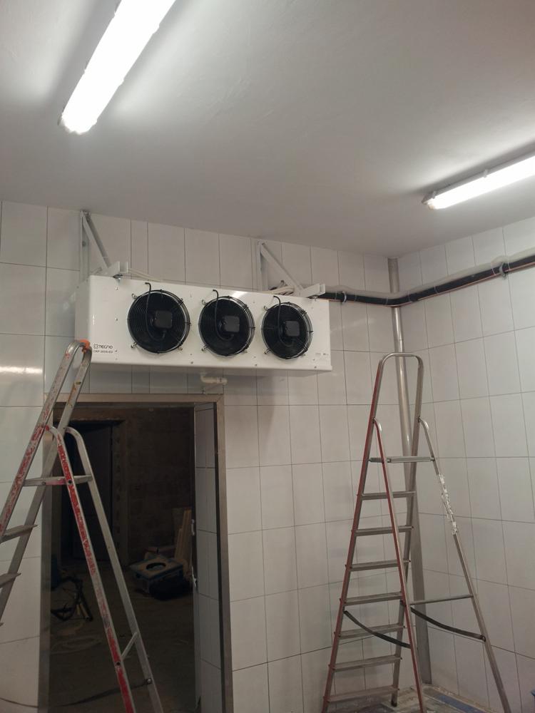 Biała chłodnica sufitowa z trzema wentylatorami zamontowana w pomieszczeniu wyłożonym białymi płytkami, widoczne dwie drabiny i otwór drzwiowy.