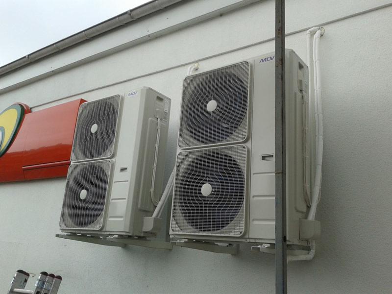 Dwa zewnętrzne moduły klimatyzacji MDV zamontowane na ścianie budynku z widocznymi przewodami i fragmentem szyldu reklamowego w tle.