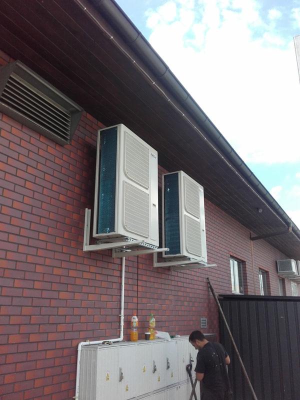 Dwa klimatyzatory zamontowane na ceglanej ścianie budynku, widoczne przewody, instalacja zewnętrzna i pracownik z narzędziem na tle pochmurnego nieba.