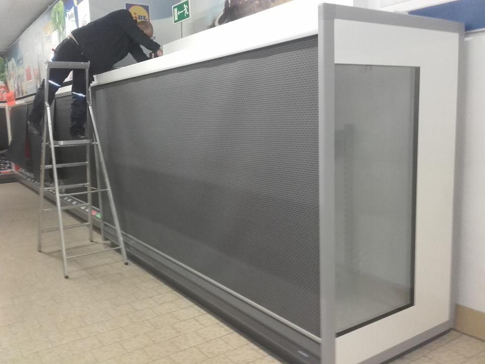 Pracownik na drabinie serwisuje chłodniczą witrynę sklepową w Kusowie, widoczny panel z perforowanej blachy i przeszklona część ekspozycyjna.