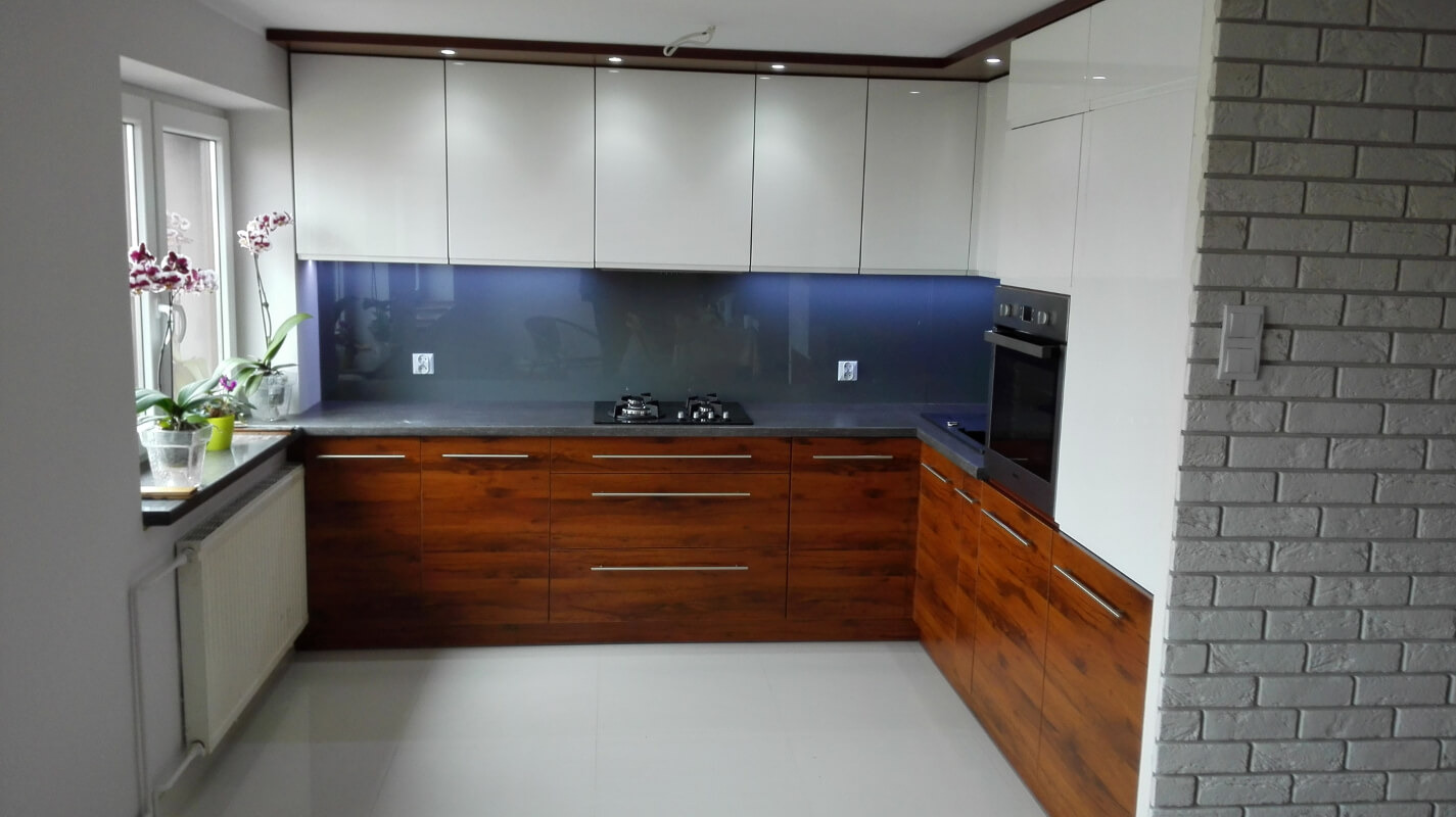 Nowoczesna kuchnia z drewnianymi frontami szafek dolnych, białymi szafkami górnymi i szarym blatem, uzupełniona o szklany panel nad blatem oraz ścianę wykończoną cegłą.