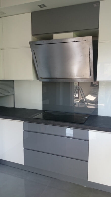 Nowoczesna kuchnia z białymi i szarymi szafkami, czarnym blatem i stalowym okapem, z odbiciem drabiny w szklanym panelu nad płytą grzewczą.