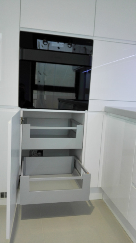 Nowoczesna, biała zabudowa kuchenna z otwartą szafką cargo z dwoma wysuwanymi półkami i piekarnikiem w zabudowie.