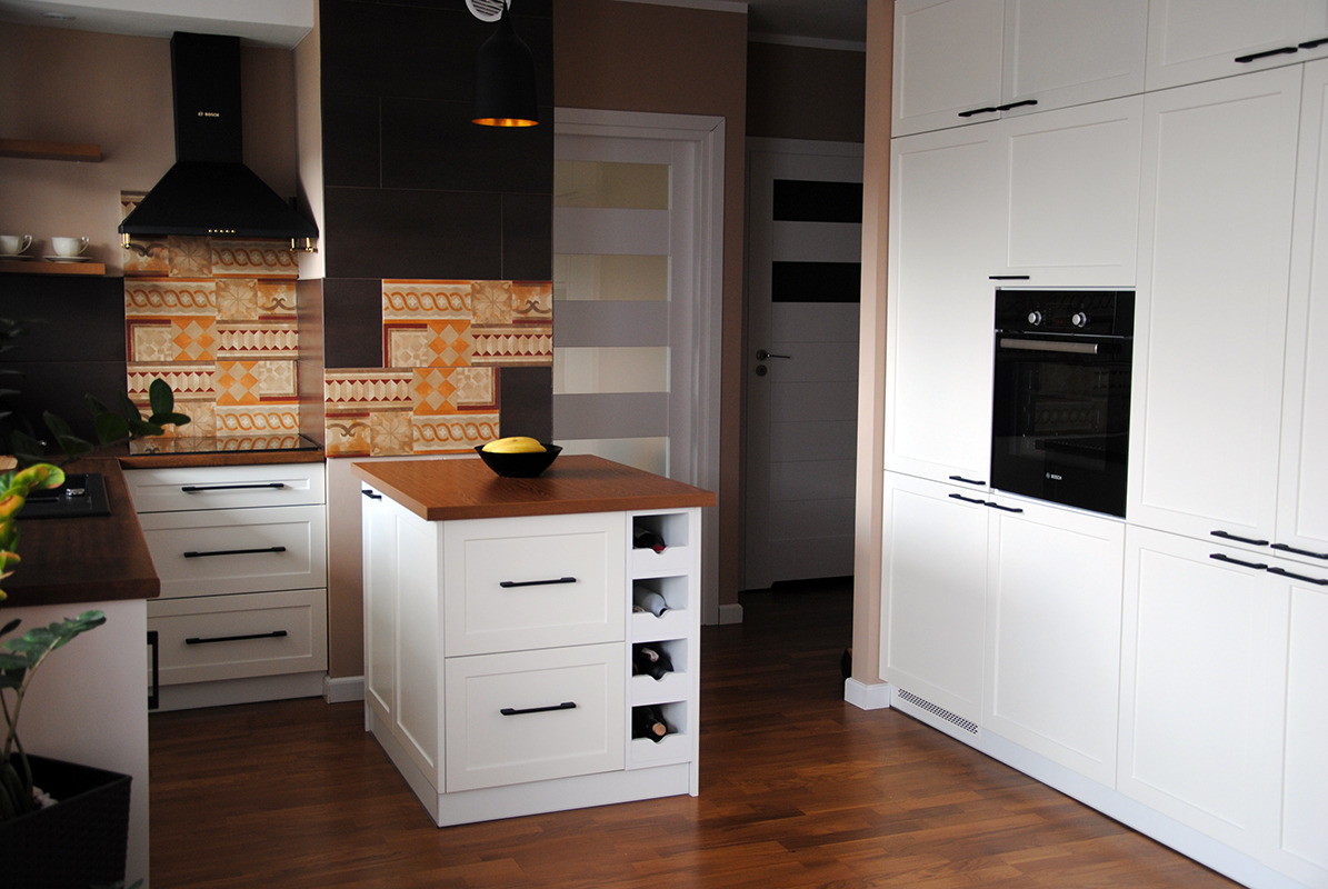 Biała zabudowa kuchenna z drewnianym blatem i wyspą z miejscem na wino, czarny okap nad płytą grzewczą, wzorzysta mozaika na ścianie.