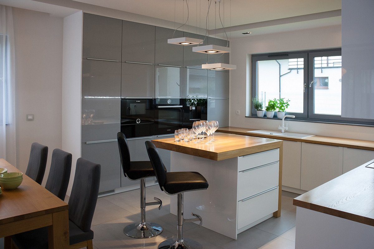 Nowoczesna kuchnia z szarymi, lakierowanymi szafkami, drewnianym blatem wyspy i minimalistycznym oświetleniem wiszącym, dodatkowo widoczne krzesła barowe i stół z krzesłami.