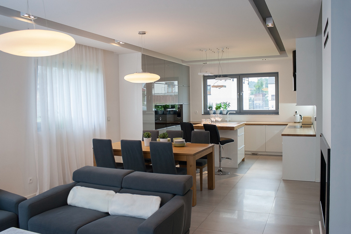 Nowoczesne wnętrze: salon z szarą sofą i drewnianym stołem jadalnym, przechodzący w jasną kuchnię z drewnianym blatem i oknem z widokiem na zieleń, oświetlone wiszącymi lampami.