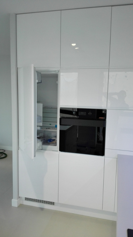 Biała zabudowa kuchenna z otwartą lodówką i wbudowanym piekarnikiem, minimalistyczny design.