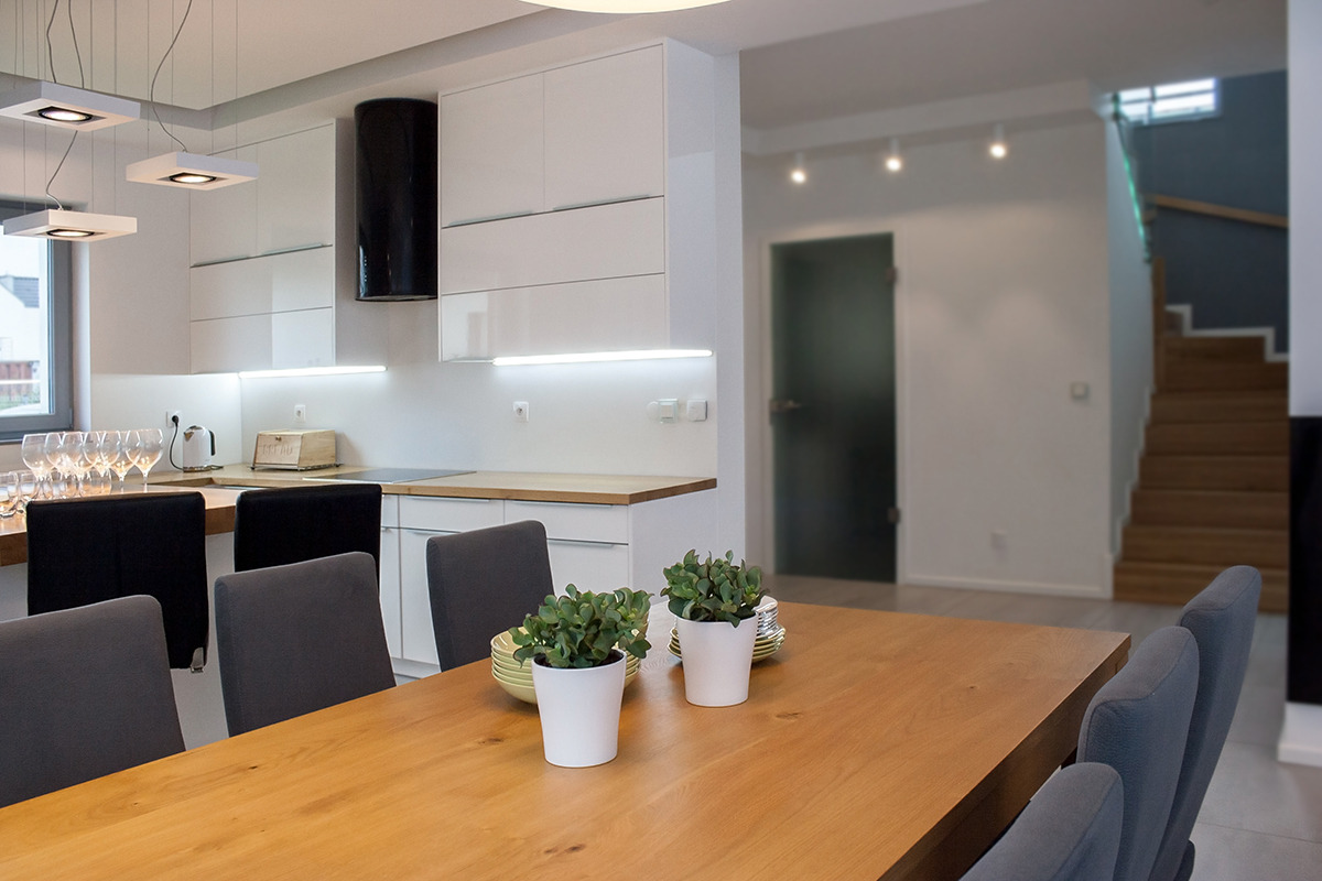 Jasna kuchnia z białymi szafkami i czarnym okapem, widok z perspektywy jadalni z drewnianym stołem i szarymi krzesłami, na stole doniczki z zielonymi roślinami.