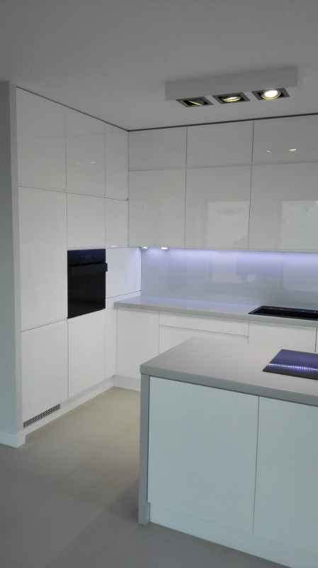 Nowoczesna, minimalistyczna kuchnia z białymi, lakierowanymi frontami, oświetleniem LED podszafkowym i wyspą kuchenną z płytą indukcyjną.