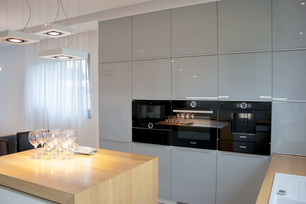 Nowoczesna kuchnia: szare, lakierowane szafki na wymiar z wbudowanymi urządzeniami Bosch (piekarnik, mikrofalówka, ekspres do kawy), drewniany blat wyspy, lampy sufitowe i ustawione kieliszki do wina.