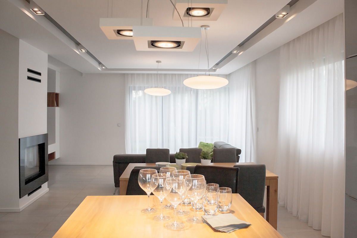 Nowoczesna jadalnia z drewnianym stołem, nakrytym szkłem i szarymi serwetkami, oświetlona designerskimi lampami sufitowymi i światłem dziennym wpadającym przez białe, zwiewne zasłony.