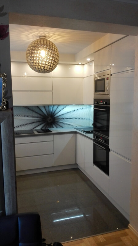 Nowoczesna, biała zabudowa kuchenna z wzorzystym panelem nad blatem, oświetlona kulistą lampą sufitową, widok z perspektywy wejścia.