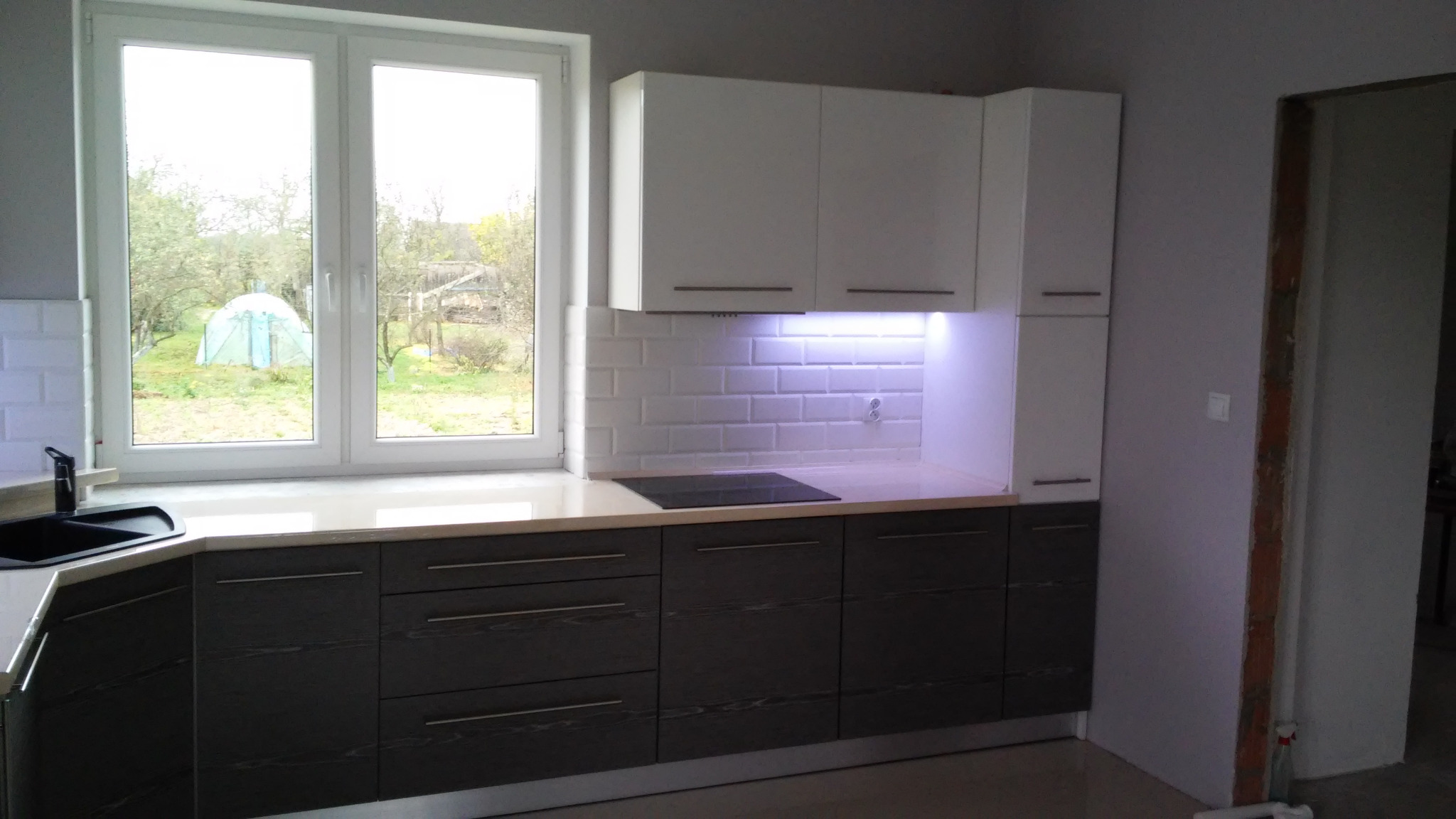Nowoczesna kuchnia z ciemnymi, drewnopodobnymi szafkami dolnymi i białymi szafkami górnymi, podświetleniem LED pod szafkami i białymi płytkami na ścianie, z widokiem na ogród przez okno.