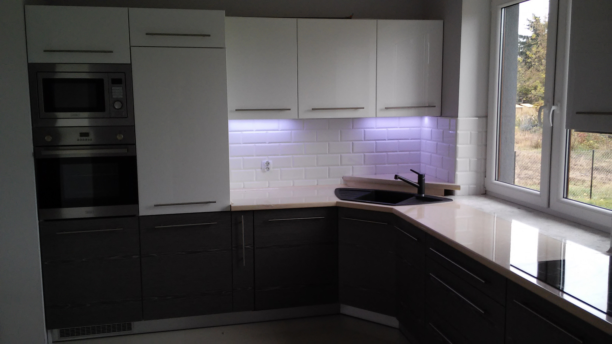 Nowoczesna kuchnia z białymi górnymi szafkami i ciemnymi dolnymi, oświetlenie LED pod szafkami, białe płytki na ścianie i czarny zlew przy oknie.