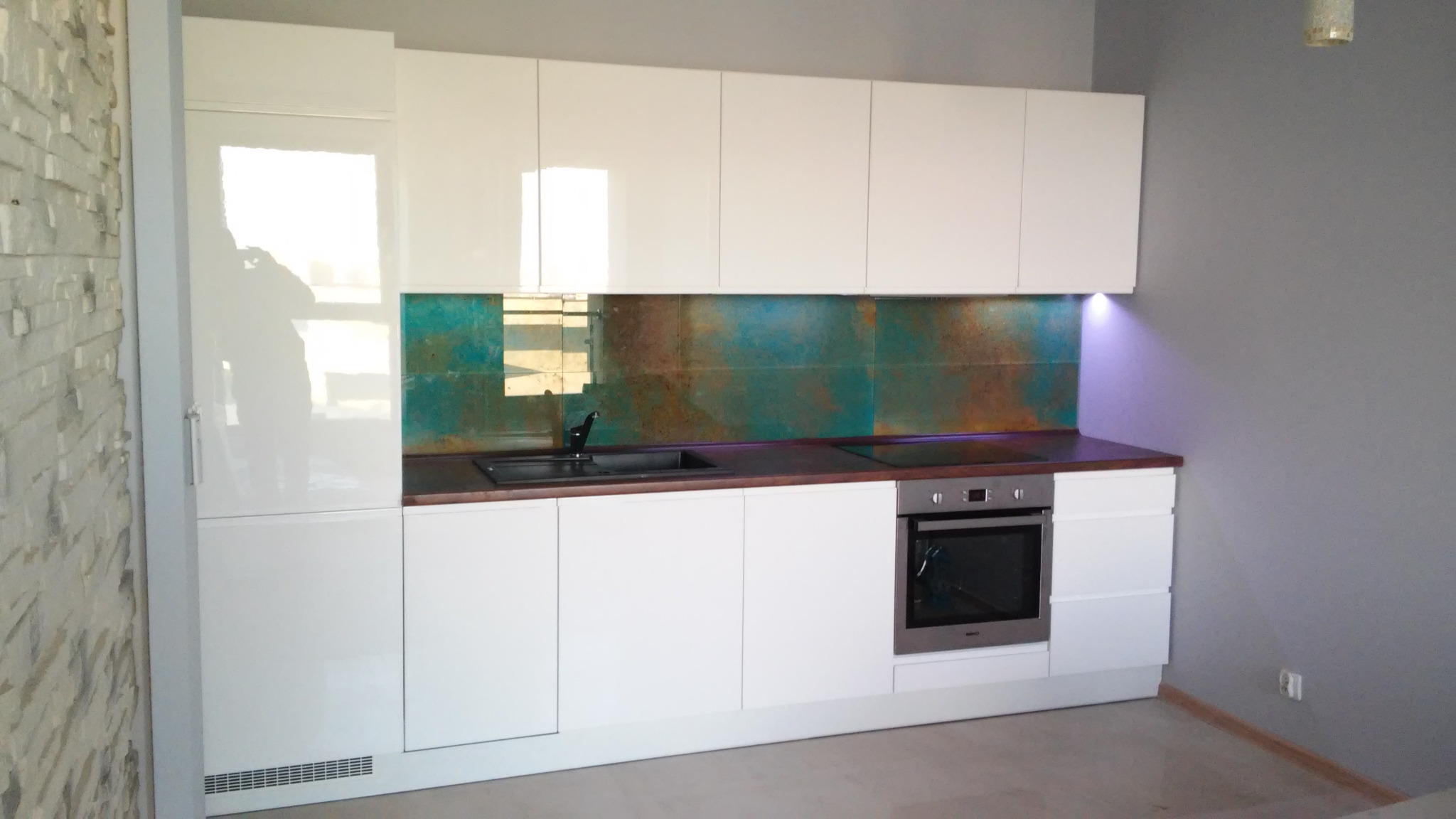 Biała zabudowa kuchenna z połyskiem, brązowy blat i dekoracyjny panel ścienny w odcieniach turkusu i brązu, oświetlenie LED pod szafkami.