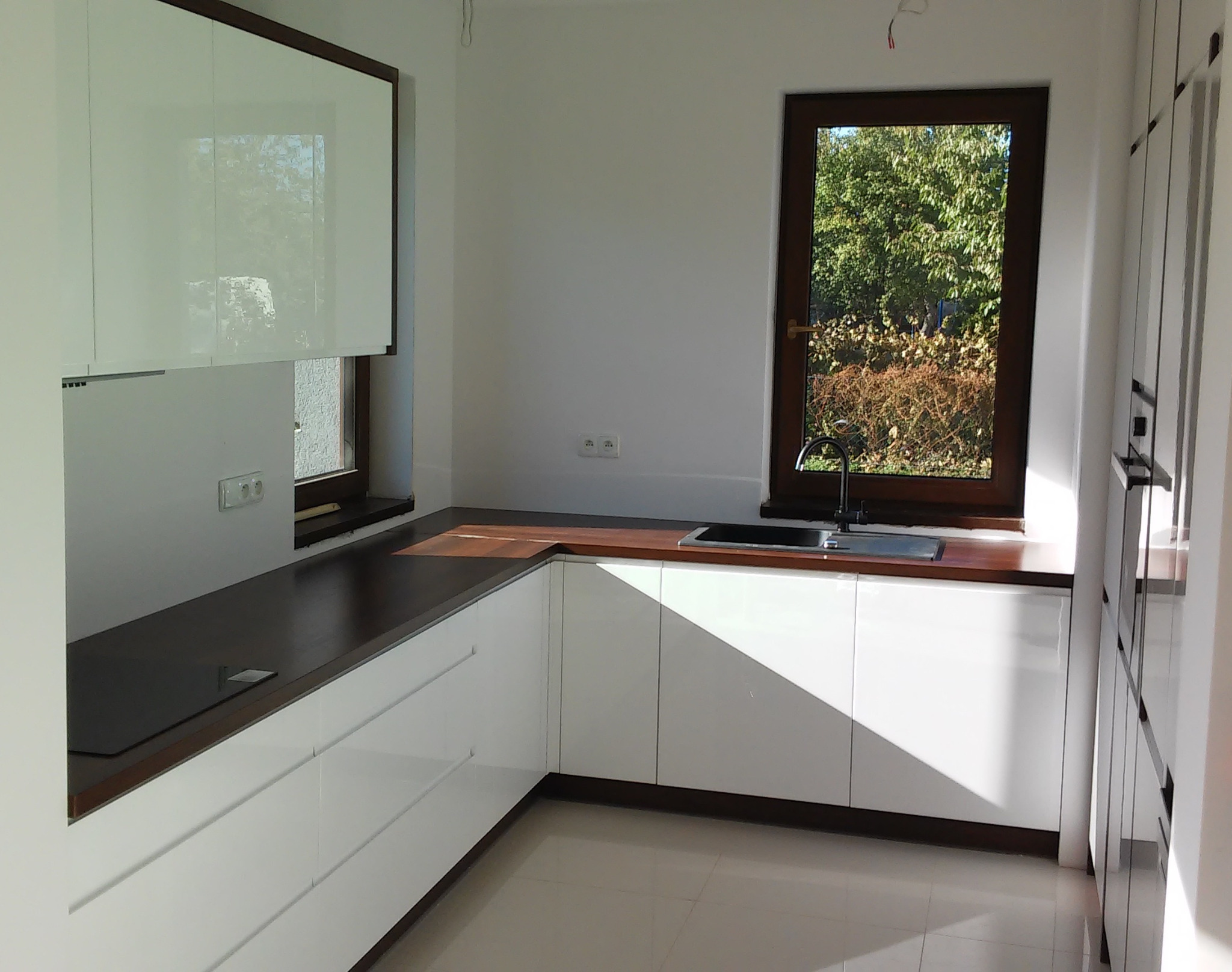 Nowoczesna kuchnia z białymi szafkami i ciemnym blatem, widok na okno z zielenią na zewnątrz.