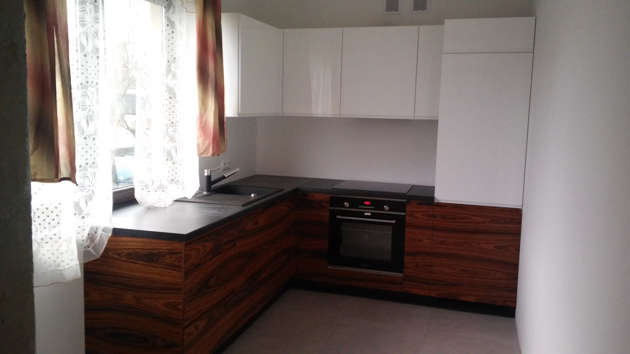 Nowoczesna kuchnia z białymi szafkami górnymi i dolnymi szafkami z ciemnego drewna o wyraźnym usłojeniu, czarny blat, widoczne okno z koronkową firanką.