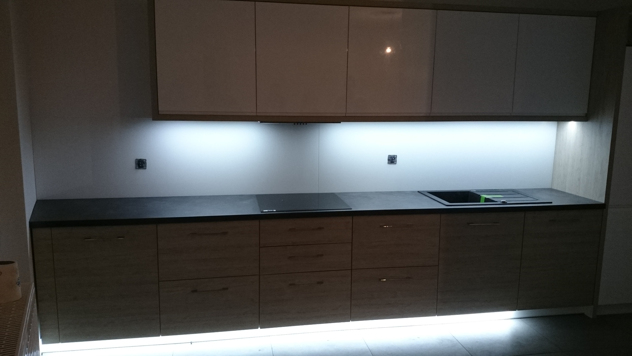 Nowoczesna zabudowa kuchenna z drewnianymi frontami szafek dolnych i białymi górnymi, podświetlana LED od dołu i od góry, z czarnym blatem i płytą indukcyjną.