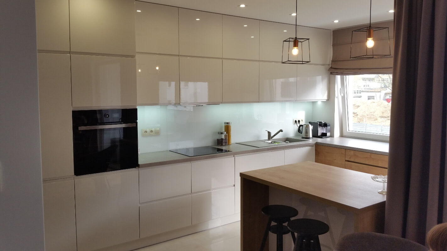 Jasna, nowoczesna kuchnia z białymi, lakierowanymi frontami, czarnym piekarnikiem w zabudowie, szklanym panelem nad blatem, wyspą z drewnianym blatem i dwoma czarnymi stołkami barowymi...