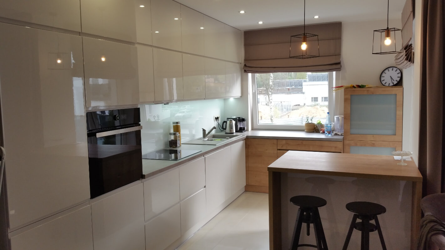 Nowoczesna, jasna kuchnia z białymi, lakierowanymi frontami szafek, drewnianym blatem wyspy i minimalistycznym oświetleniem wiszącym.