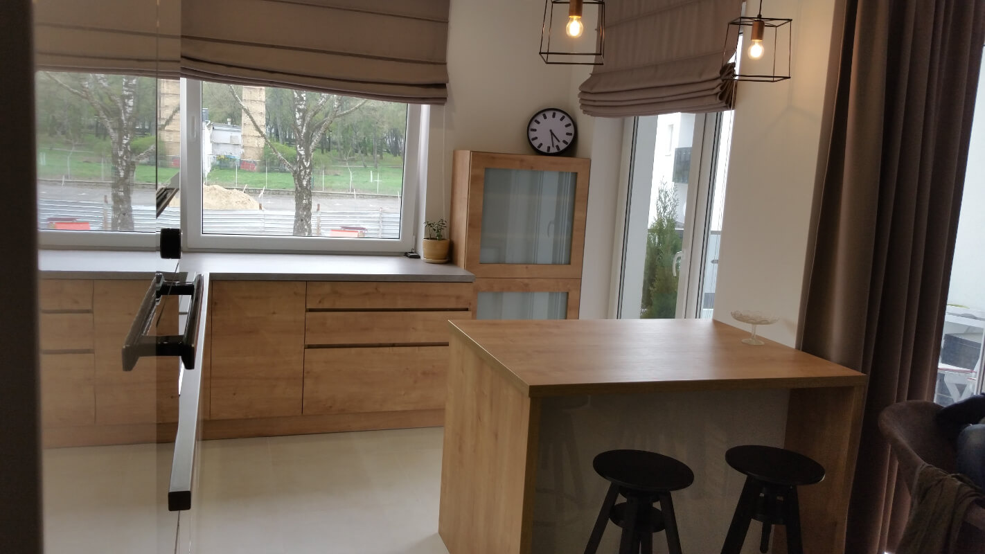 Jasna kuchnia z drewnianymi szafkami, wyspą z hokerami i oknem z widokiem na zewnątrz, oświetlona wiszącymi lampami.