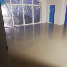 Wnętrze domu w trakcie prac wykończeniowych: świeżo wylana, samopoziomująca posadzka, okna i drzwi zabezpieczone folią malarską, widoczne elementy konstrukcji dachu.