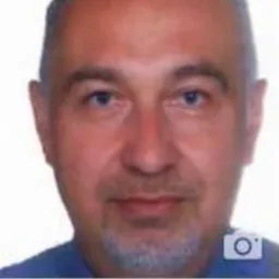 Portret mężczyzny w niebieskiej koszuli, z krótkimi, ciemnymi włosami i siwą brodą, na białym tle.