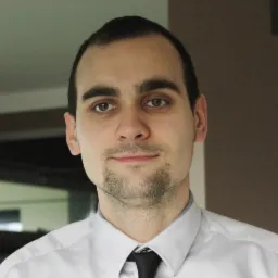 Portret mężczyzny w białej koszuli i czarnym krawacie, z ciemnymi włosami i zarostem, patrzącego prosto w obiektyw.