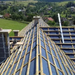 Konstrukcja dachu z drewnianymi łatami na folii, komin z szarych bloczków, poziomica pionowo oparta o konstrukcję, wiadro na drewnianej desce, widok na wieś w tle.