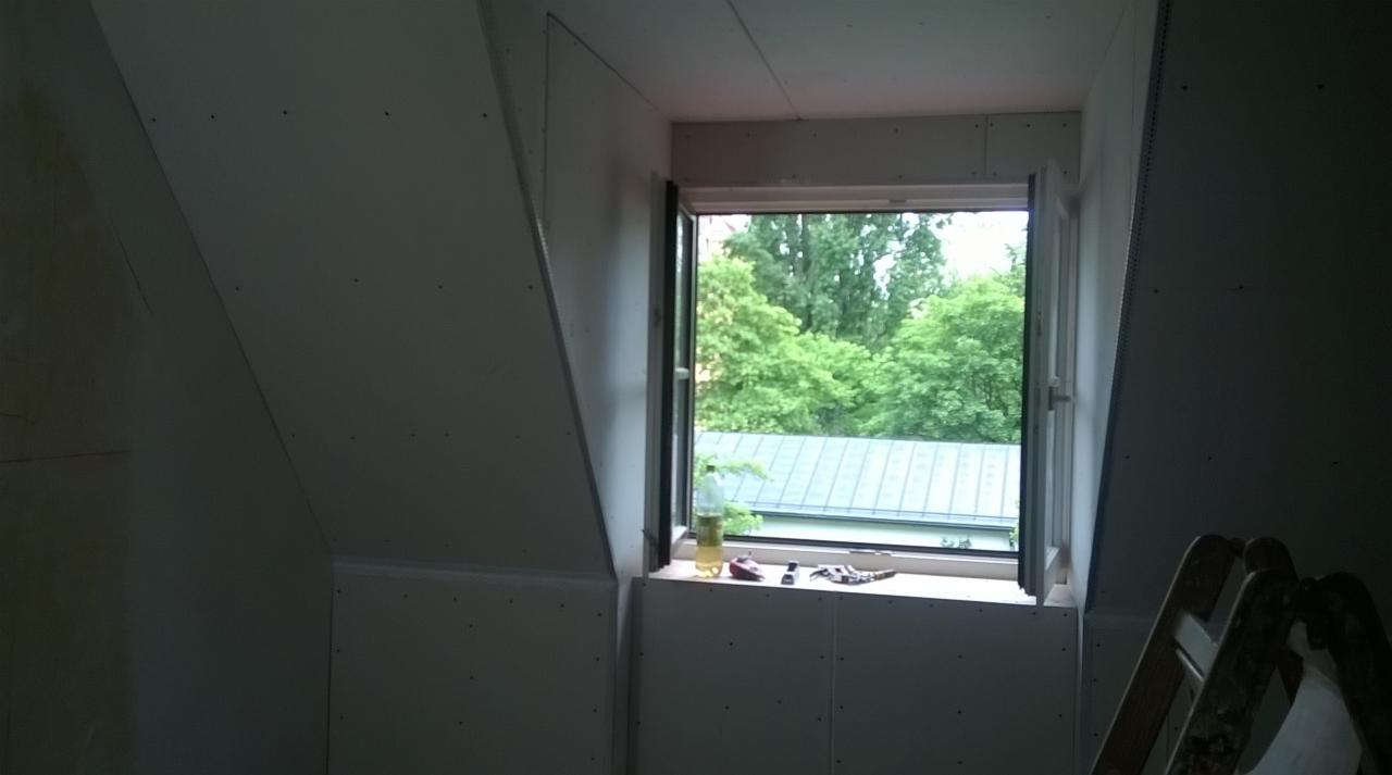 Poddasze w trakcie wykańczania płytami kartonowo-gipsowymi, widoczne narzędzia na parapecie okna dachowego z widokiem na zieleń.