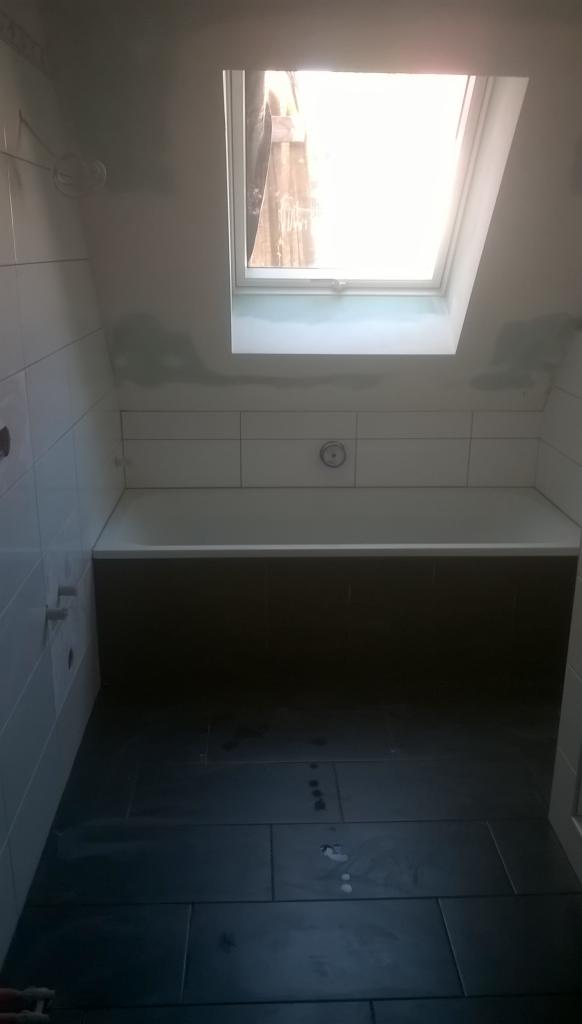 Wnętrze łazienki w trakcie prac wykończeniowych; widoczna wanna obudowana ciemnymi płytkami, białe płytki na ścianach, czarna podłoga z płytek, okno dachowe w skosie; ślady zaprawy na ścianach...