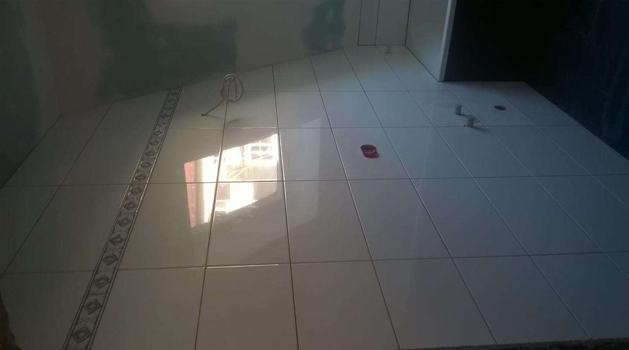 Świeżo ułożone, białe, błyszczące płytki podłogowe w łazience, z dekoracyjnym pasem płytek przy ścianie, widoczne otwory na instalacje i narzędzie do mieszania zaprawy.