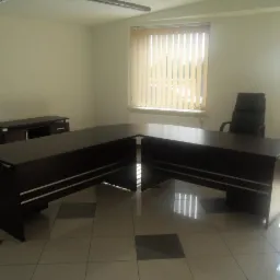 Duże, ciemne biurko w kształcie litery U z białymi akcentami, czarne krzesło biurowe na kółkach, szafka biurowa i okna z pionowymi żaluzjami w jasnym biurze z kafelkową podłogą.