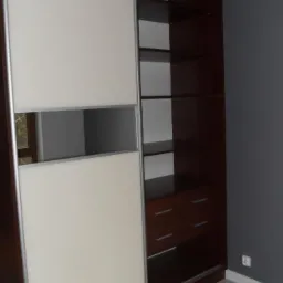 Wnętrze szafy przesuwnej z kremowymi drzwiami z poziomym przeszkleniem i otwartą częścią z ciemnego drewna z półkami i szufladami na tle szarej ściany.