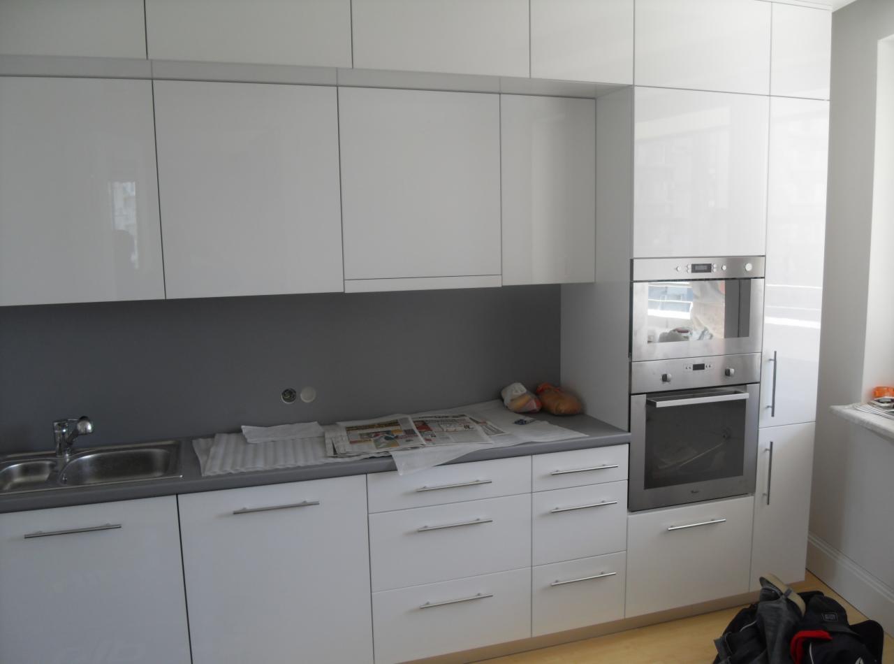 Nowoczesna biała kuchnia z błyszczącymi frontami, szarym blatem i zabudowanym piekarnikiem, na blacie leżą gazety i torby foliowe, widoczne niedoskonałości montażu.
