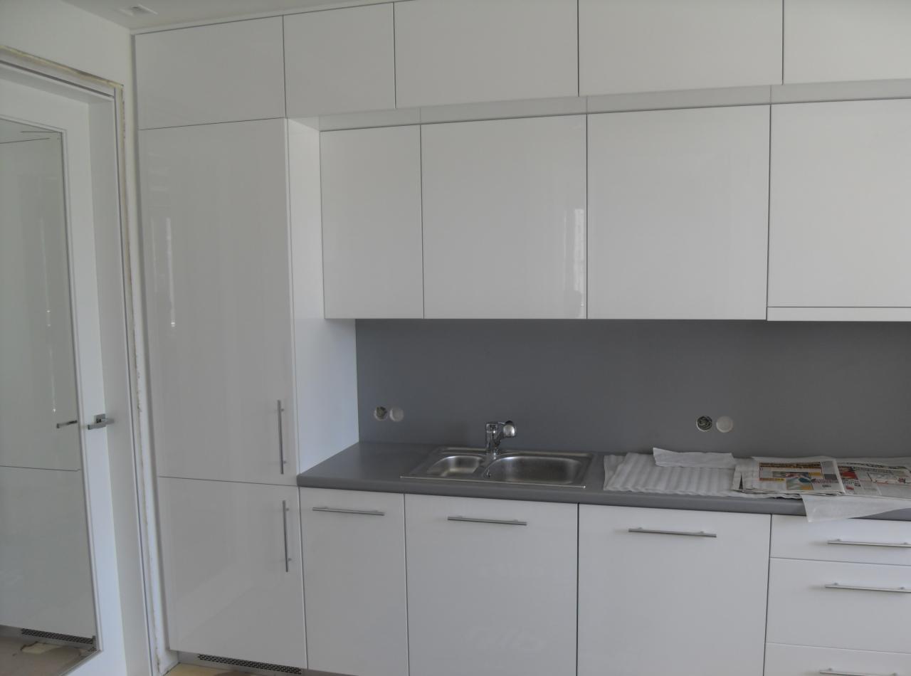 Biała, minimalistyczna kuchnia z błyszczącymi frontami szafek, szarym blatem i zlewozmywakiem dwukomorowym, uzupełniona gazetami leżącymi na blacie obok zlewu.