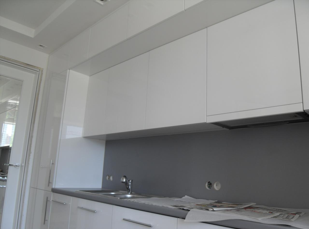 Białe, lakierowane szafki kuchenne wiszące, z szarym panelem ściennym, widoczne gniazdka elektryczne i gazeta na blacie, jeszcze przed zakończeniem montażu.