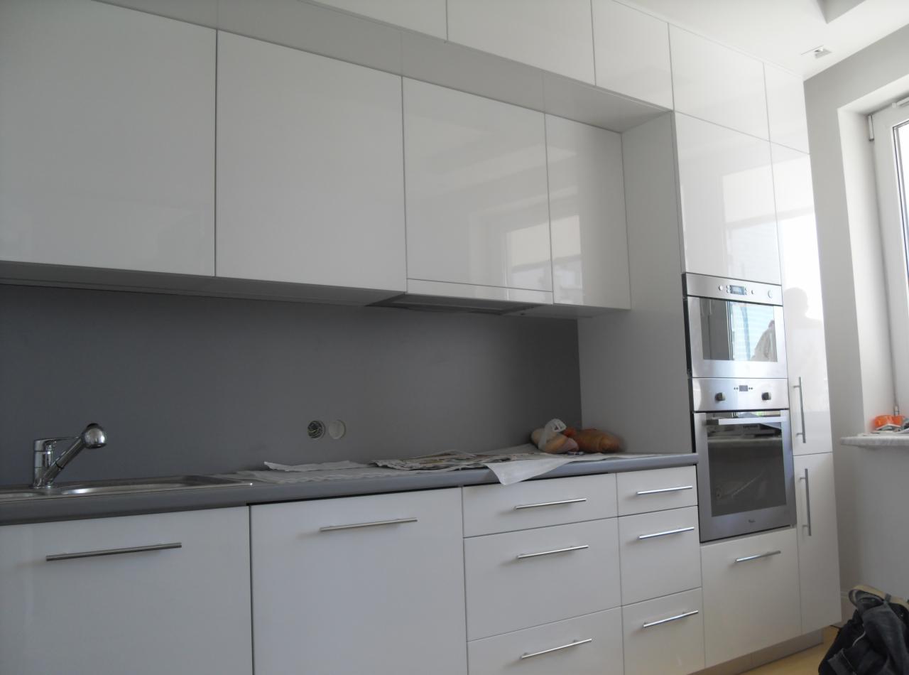 Nowoczesna, biała kuchnia z połyskiem, widoczne szafki górne i dolne, zlewozmywak, piekarnik w zabudowie oraz przygotowane miejsce do pracy na blacie.