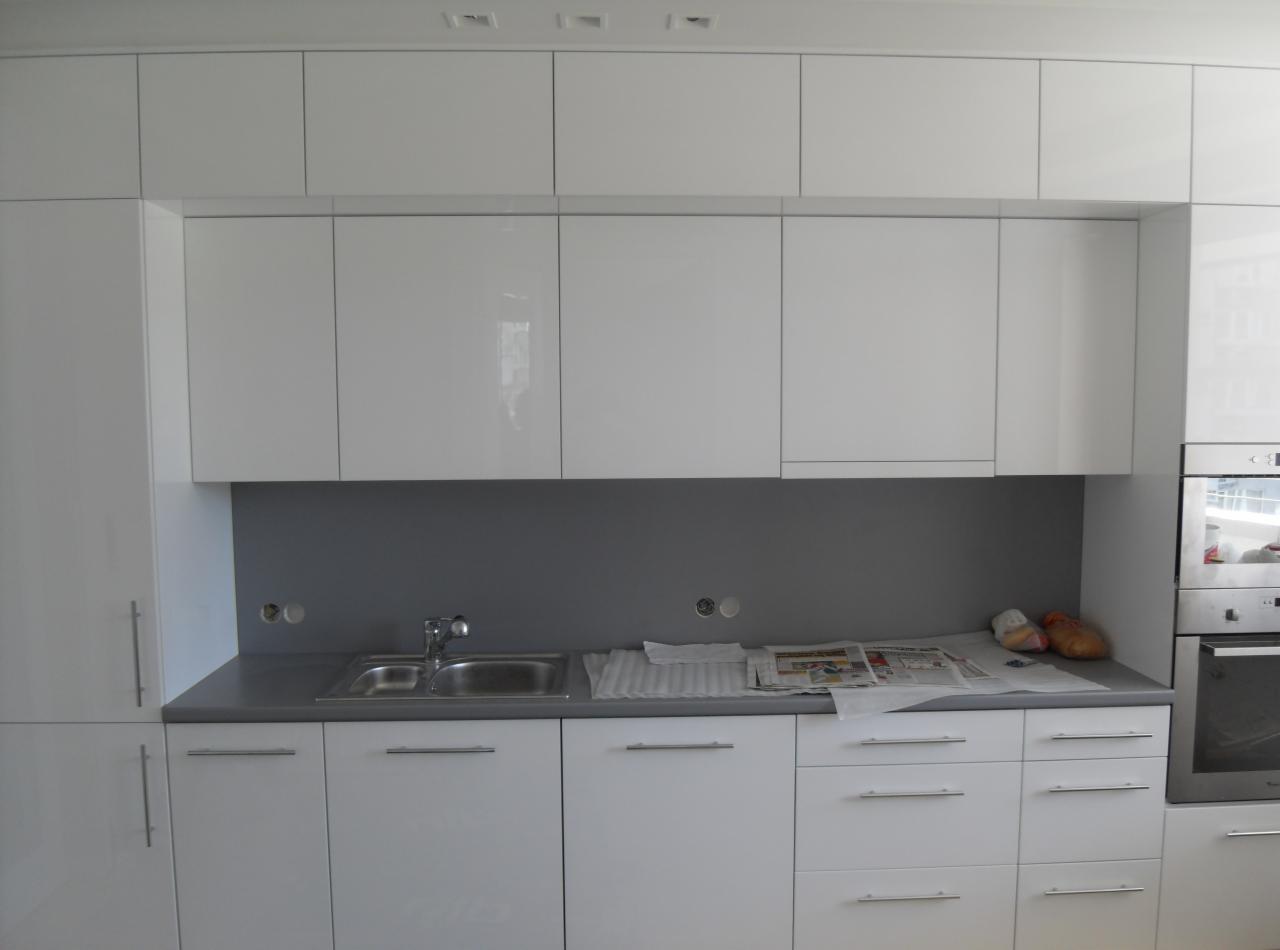 Biała, nowoczesna zabudowa kuchenna z błyszczącymi frontami, szarym blatem i płytkami, widoczny zlew, bateria, gazeta i otwory na gniazdka elektryczne w ścianie.