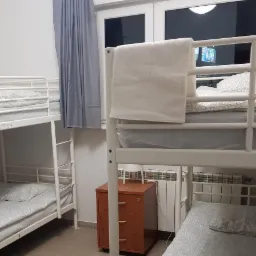 Pokój wieloosobowy z trzema łóżkami piętrowymi z białą ramą, szarą pościelą w drobne wzory i brązową komodą z trzema szufladami.