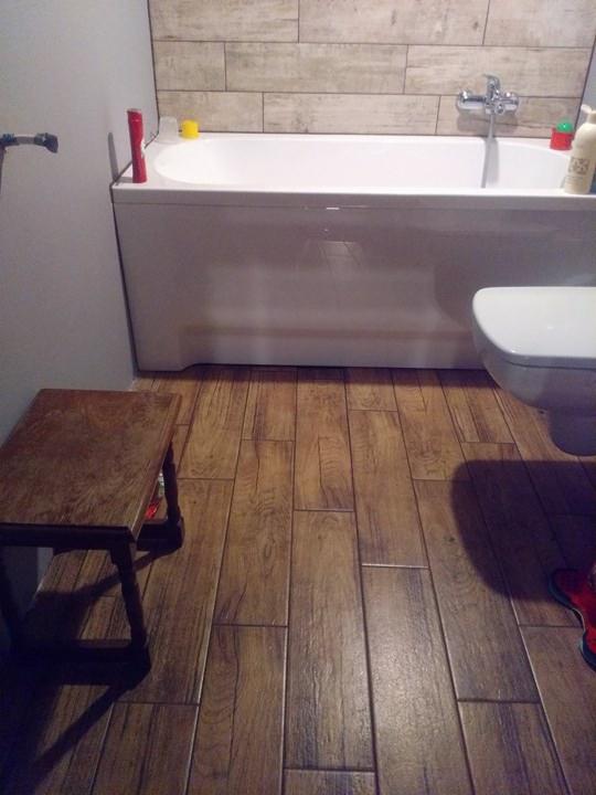 Łazienka po remoncie: płytki imitujące drewno na podłodze i ścianie nad wanną, biała wanna i toaleta, mały drewniany taboret przy ścianie.