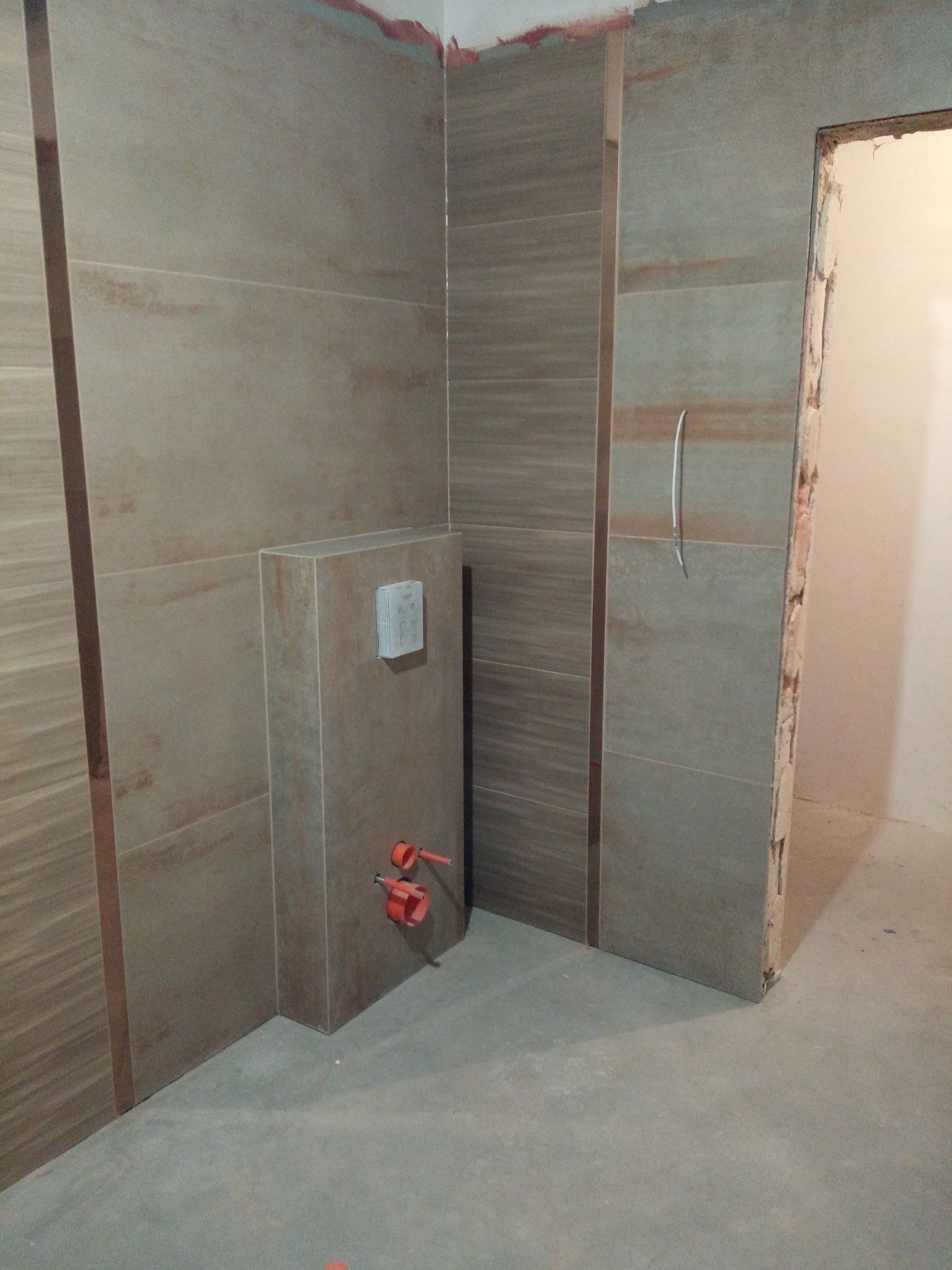 Ściana łazienki wyłożona płytkami imitującymi drewno, widoczny stelaż podtynkowy z przyłączami wodnymi i częściowo wykończone obramowanie drzwi.