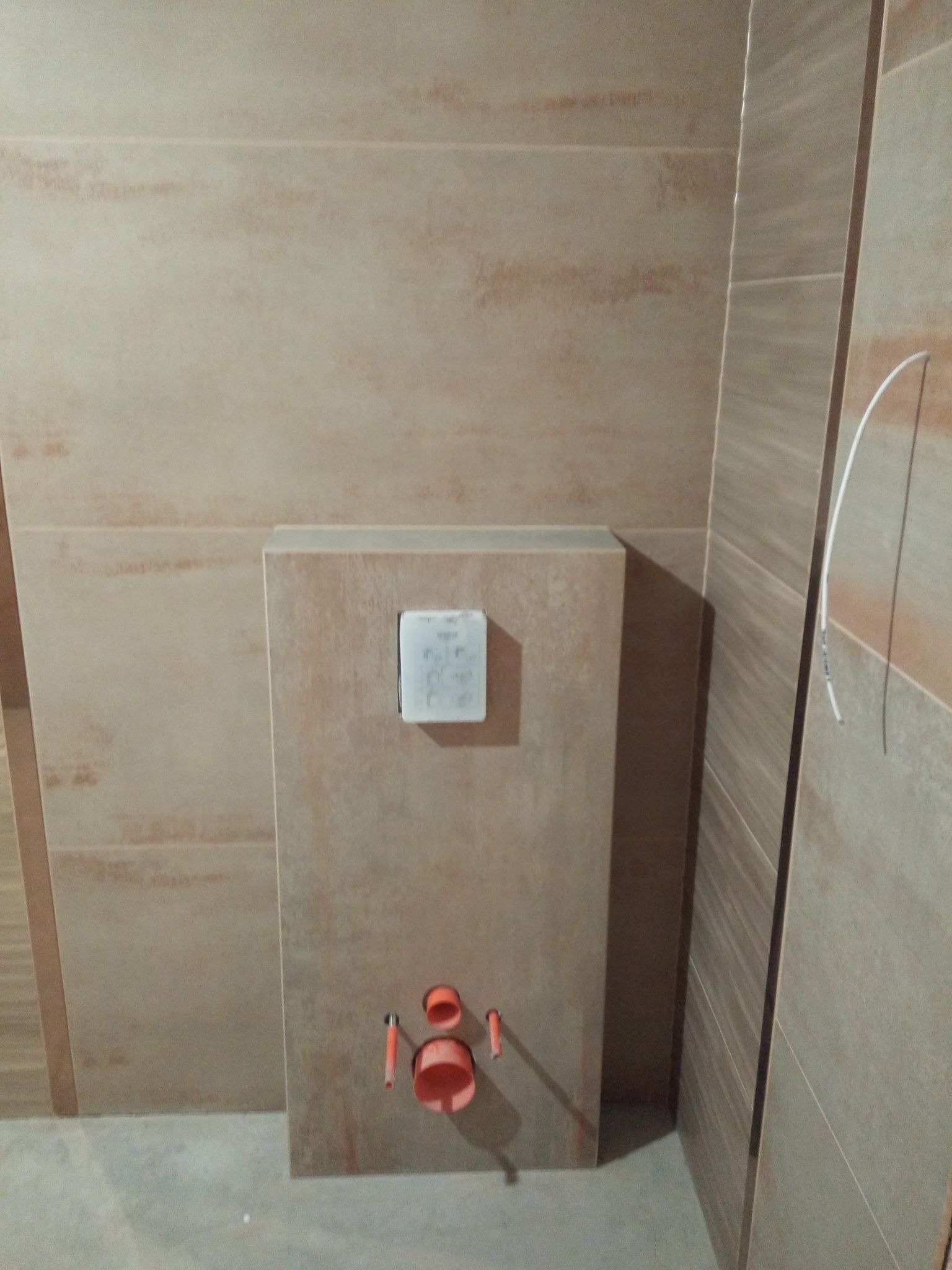 Ściana łazienki wyłożona beżowymi płytkami imitującymi drewno, z zamontowaną podtynkową instalacją sanitarną i przyciskiem spłukującym, widoczne fragmenty rur.