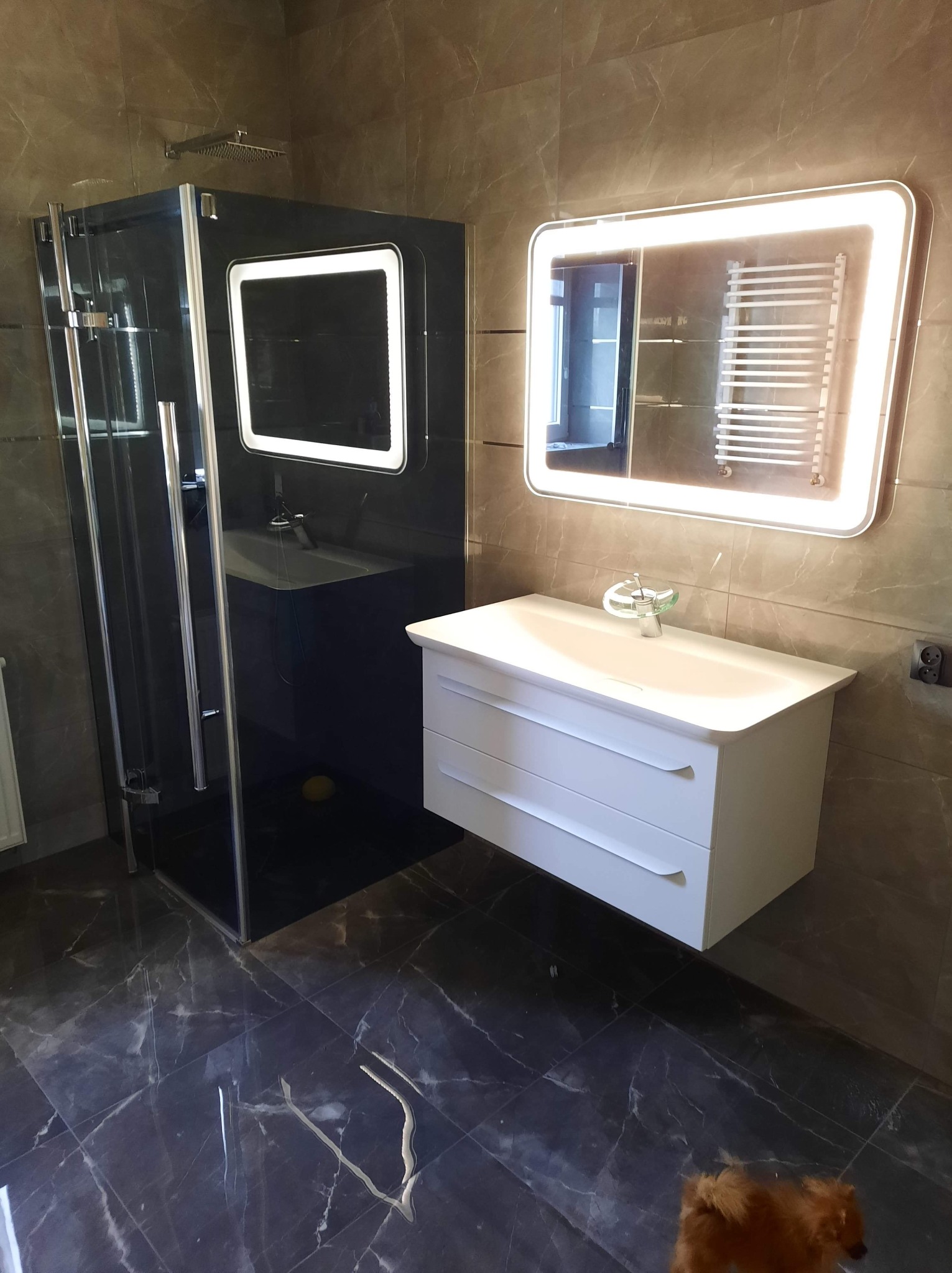 Nowoczesna łazienka z czarną kabiną prysznicową, białym umywalką z szafką i podświetlanym lustrem, wykończona płytkami imitującymi marmur; mały pies w rogu kadru.