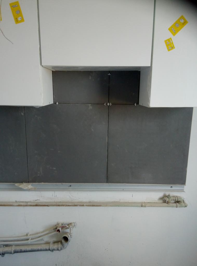 Montaż ciemnych płytek na ścianie nad blatem w kuchni, widoczne krzyżyki dystansowe, mocowania i rury instalacyjne.