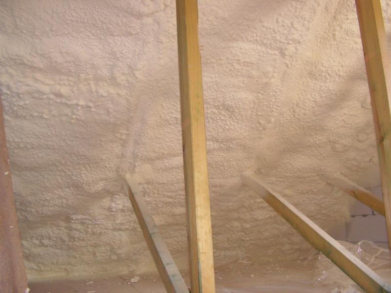 Świeżo nałożona, biała pianka izolacyjna wypełnia przestrzeń między drewnianymi krokwiami poddasza, widoczna folia ochronna na podłodze.