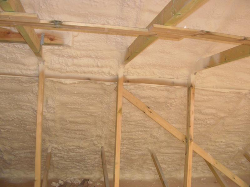 Poddasze z drewnianą konstrukcją pokryte grubą warstwą jasnej piany izolacyjnej, widoczne nierówności i struktura piany.