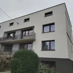 Dwupiętrowy budynek z jasną elewacją i ciemnoszarą podmurówką, widok z dołu na balkon z metalową balustradą i nowoczesne okna w brązowych ramach.