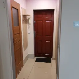 Przedpokój z brązowymi drzwiami wejściowymi, drewnianą szafką z wieszakami, kremowymi płytkami podłogowymi i oświetleniem LED na suficie.